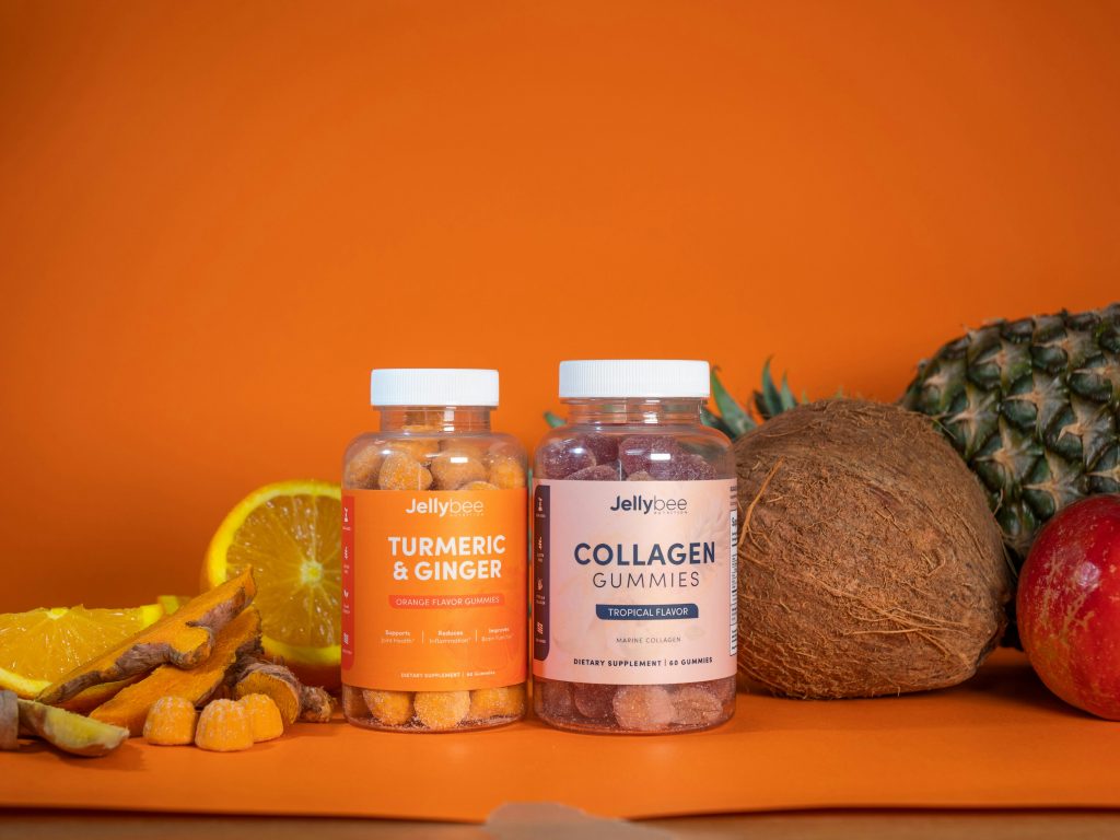 Two bottles of gummy supplements labeled “Turmeric & Ginger” and “Collagen Gummies” are placed on an orange background, surrounded by fresh fruits like orange slices, pineapple, coconut, and an apple.