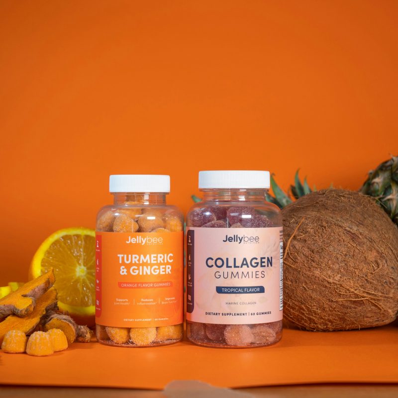 Two bottles of gummy supplements labeled “Turmeric & Ginger” and “Collagen Gummies” are placed on an orange background, surrounded by fresh fruits like orange slices, pineapple, coconut, and an apple.